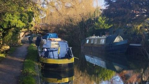 Towpath on left with line of canal boats on both sides of water with bridge seen behind and trees arching over the water - it is a bright, sunny day.