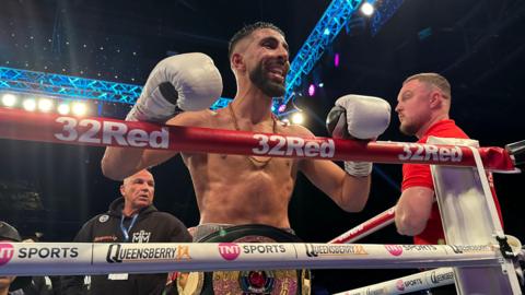 Shabaz Masoud, a boxer wearing white gloves, smiles as he is stood in a boxing ring with two other men also stood in the ring with him. He is wearing a large boxing title belt.