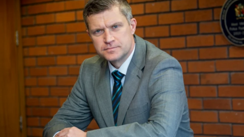 Cooke is wearing a grey suit and tie and white shirt inside a building. There is a brock wall behind him and he is leaning on a banister rail and looking at the camera.