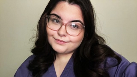Ayla Jones is a white woman with long dark hair and large light brown framed glasses. She is wearing a purple medical scrubs top and is looking slightly to the right of the picture and smiling.