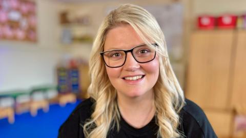 A woman with long blonde hair, smiling at the camera and wearing dark-rimmed glasses and and a black top.