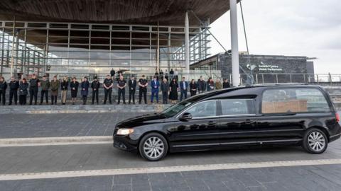 A black funeral car with a flower-topped wicker casket inside. It is driving in front of the grey building of the Welsh parliament, with a line of people dressed in smart clothes outside it, all with their heads bowed.