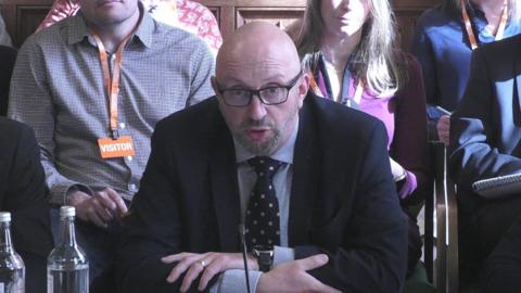 David Hinton in a suit speaks to a select committee
