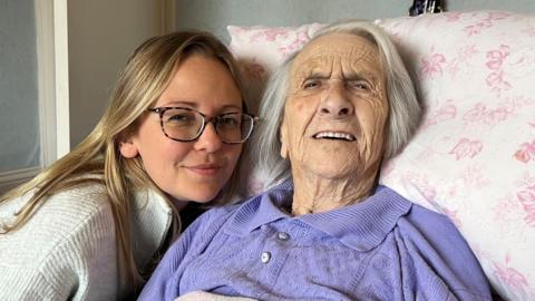 A woman in her thirties is kneeling next to the bed of her 98-year old grandmother. The younger woman is leaning in towards the older woman for the photograph. 