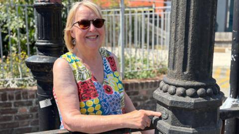 A woman in a patterned, floral dress smiles at the camera. She has blonde hair and is wearing sunglasses. She is pointing at a cast iron pipe.