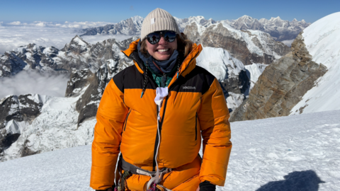 Karolina Pakenaite on a mountain side. She is wearing a bright orange puffer coat with black shoulders. She has on a light brown beanie hat and dark sunglasses. The background behind her is the view of mountain peaks with snowy white tips and a pale blue sky.