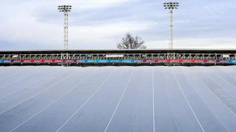 A covering over the top of Luton Town Football Club's pitch, it is white and very large with lines across it. A football stand can be seen in the distance, with a free behind it and two large flood lights.