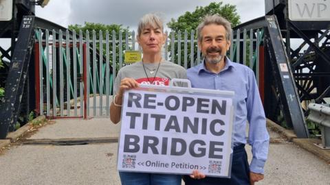 A man with short hair wearing a blue shirt and blue trousers, and a woman with short hair wearing a grey T-shirt with a red and white logo and blue jeans. She is holding a sign reading: 'RE-OPEN TITANTIC BRIDGE. ONLINE PETITION'.