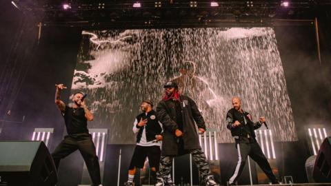 The four members of JLS on stage singing. There is a screen behind them playing a music video with rain pouring down. They are in black clothes and have handheld black microphones