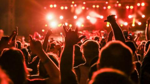 A band playing on stage is shown in the background but out of focus with bright red lights, with the back of people's heads shown in the foreground as some raise their hands in the air in celebration.