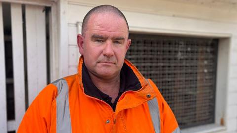 Pigeon fancier Mick Ready standing in front of a pigeon loft on his allotment, wearing an orange hi-viz coat with florescent stripes.