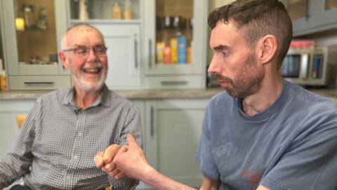 David Sincock (left) sitting next to his dad, Ian, in a kitchen. Ian is smiling at him as they bump fists. Ian has short grey hair and a beard and is wearing glasses along with a grey checked shirt. David has short dark hair and a beard. He is wearing a blue T-shirt. Behind them are cupboards filled with items of food, and a microwave can be seen on a worktop over David's shoulder.