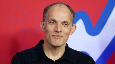 Tuchel smiling with a bright red background behind him. He's wearing a black polo shirt.