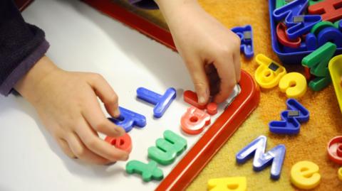 A child's hands are putting green, red and blue plastic letters on a white magnet board. More letters are scattered on the carpet next to the board.
