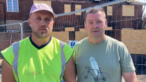 Mike has a high viz shirt on and a cap. He has a beard. Roy is stood next to him in a t-shirt and glasses on his head. He has short brown hair. They are stood in front of their damaged home which are boarded up with a metal railing surrounding them.