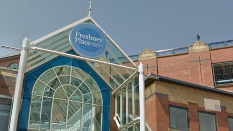 The glass canopied Entrance to Freshney Place with a blue name sign above the door and the red brick multi-storey car park in the background