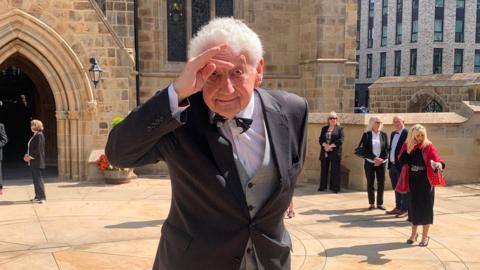 A man with grey hair makes a hand salute to his head outside a cathedral