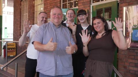 Five people are standing outside a building with their thumbs up or hands waving for the camera.