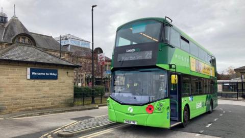 A double decker bus in light and green livery parked next a stone building with a sign that reads 'welcome to Ilkley' on its side.