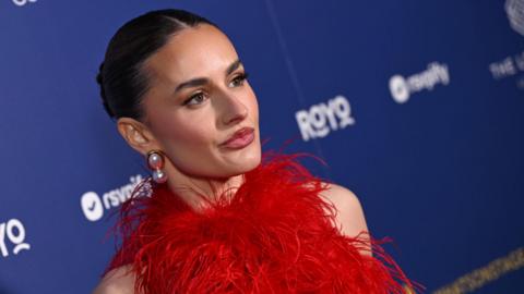 A zoomed in, slanted angle of Amber Davies, in a red feathered dress and wearing big pearl earrings. She stands in front of a branded blue wall with sponsor logos. Her dark hair is pulled back into a slick bun. 