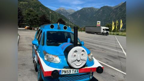 The four-door car with Thomas the Tank Engine frontage. The car has been sprayed blue with red stripes. Thomas's smiling face is stuck to the front with a funnel poking out the top and imitation wheels have been stuck to the sides of the car to complete the look. He is in a car park in the Alps and huge tree-covered mountains rise behind him.