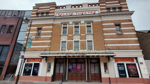 Exterior shot of Watford Palace Theatre taken in the day. It is an art deco building, with shades of brown. The sign reads: Palace Theatre.