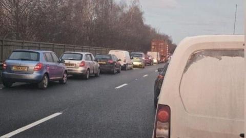 Queueing traffic on a dual carriageway road. A row of queuing vehicles can be seen in one lane while the back of a white van is nearest the viewer. A wooden fence and trees are beyond the queuing traffic