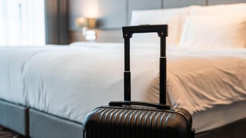 A small, black hard cased suitcase with its handle up is positioned next to a made bed in a hotel room.