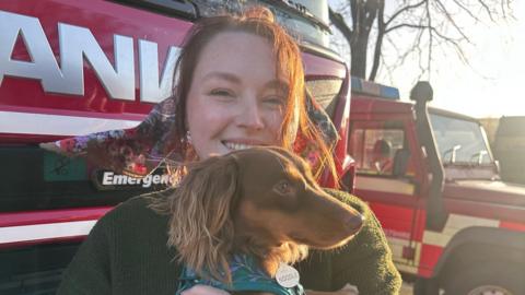 Image of a woman who is smiling at the camera while holding a small brown dog. Her hair is tied up in a pony tail with a floral scarf. In the background can be seen a number of fire service vehicles.