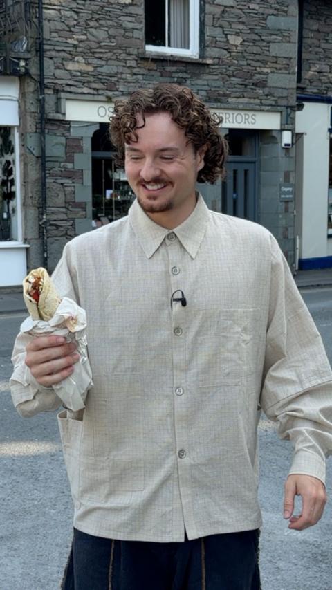 A person with curly hair and a light beige button-up shirt smiles while holding a wrapped kebab on a street in Grasmere, with traditional stone buildings in the background