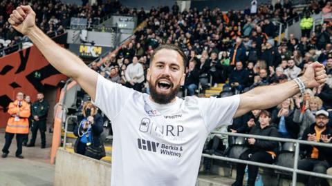 Mike has a think brown beard and short-hair the same colour. He is euphoric with both his hands outstretched running in a packed stadium.