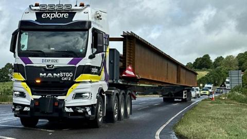 A lorry with a large load, looking like a metal beam, driving along a road with vehicles behind it. A grass verge is on the right and there are trees in the distance.