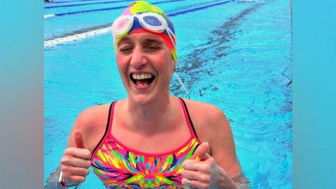 Melanie Barratt is in a blue swimming pool. She is wearing a swimming costume that is colourful tie-dye pattern, with pink, yellow, green, orange, blue and purple. She is holding both thumbs up and smiling. She is wearing a matching tie-dye swimming cap on her head, and has white goggles on her head