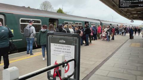 Dozens of people stood on a train platform waiting to board a dark green Great Western Railway train. 