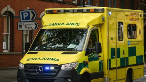 A yellow ambulance with emergency lights on. It has yellow and green squares along the side and ambulance is written in green letters. 