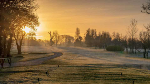A general view picture of the golf course on a pleasant morning, with the sun rising behind trees on the left of the picture. 