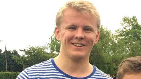 A blond-haired teenager in a blue and white striped shirt looks into the camera with trees in the background