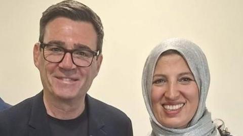 A man in a dark suit and T shirt, with dark-framed glasses, stands with a woman wearing a silver headscarf.