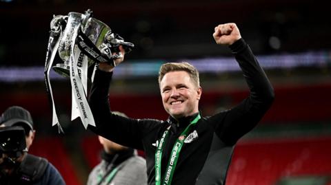 Eddie Howe holding the Carabao Cup aloft at Wembley. He has short fair hair and stubble and is wearing a black Newcastle United top. He is holding the silver trophy in his right hand. It is decorated with black-and-white ribbons.