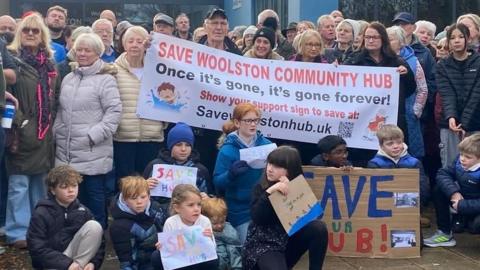 Large crowd of people at a protest with 10 children crouching in front on the ground holding some crayon-drawn signs saying "save the hub". The adults stand behind with a large banner saying "Save Woolston Community Hub - Once it's gone, it's gone forever".