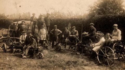 A sepia-tinged photograph of a large group of men sitting and standing in a field around farming machinery, including an early tractor. 