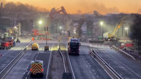 The eight lanes of the motorway are closed while multiple cranes work to pull down the concrete bridge overhead. Artificial work lights illuminate the dust rising from the works as the area is cordoned off behind plastic barriers. Vehicles are parked in front including two red and yellow highways' vans and a lorry.
