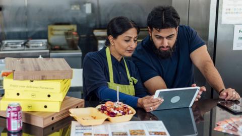 Stock photo shows two business owners looking at a tablet at the counter of a takeaway restaurant with food boxes on the counter and cooking appliances in the background.
