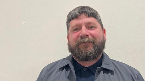 Jeff Taylor smiles at the camera as he stands in front of a white wall. He is wearing a grey coat and blue button up shirt. He has a beard and grey hair.