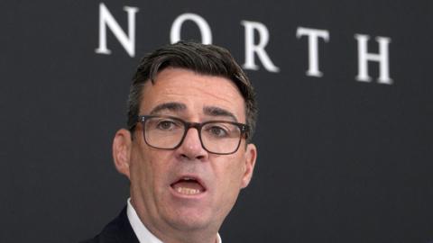 A man with black hair and glasses stands in front of a sign saying 'Number 10 of the North'.