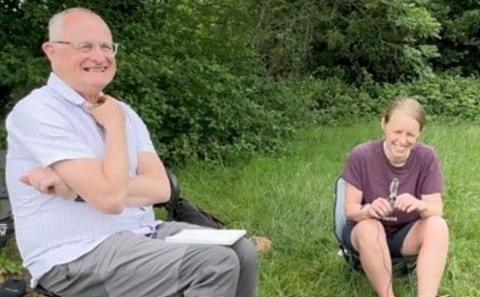 A man and a woman sitting on chairs in a field with hedges in the background