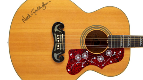 A brown acoustic guitar is pictured, with Noel Gallagher's signature seen on the body of the guitar. Around the sound hole is a detailed red pattern.