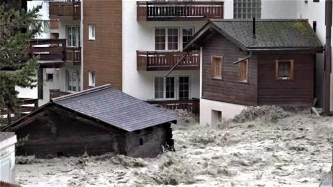 Buildings are submerged in brown floodwater, which splashes with the churning current