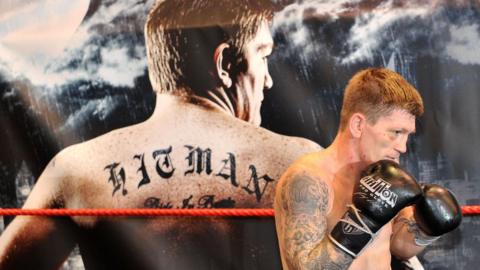A wide shot of Ricky Hatton wearing black boxing gloves. In the background is an image of Hatton's back, with the tattoo 'hitman' written across it.