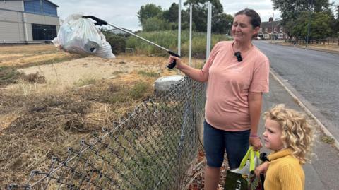 A woman in a pink t-shirt holds up a bag of litter with a litter picker as her young daughter looks on 